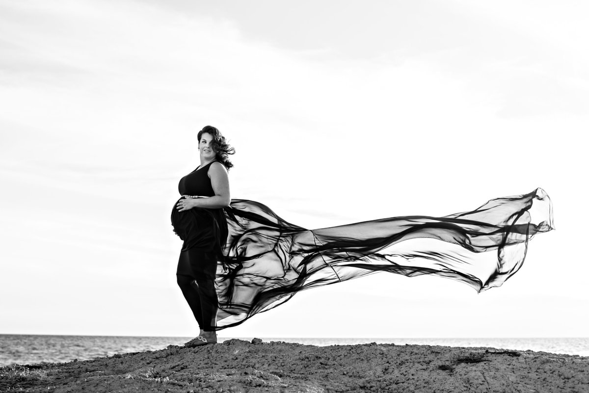 A mother to be holds her pregnant belly and her dress blows in the wind at the beach.