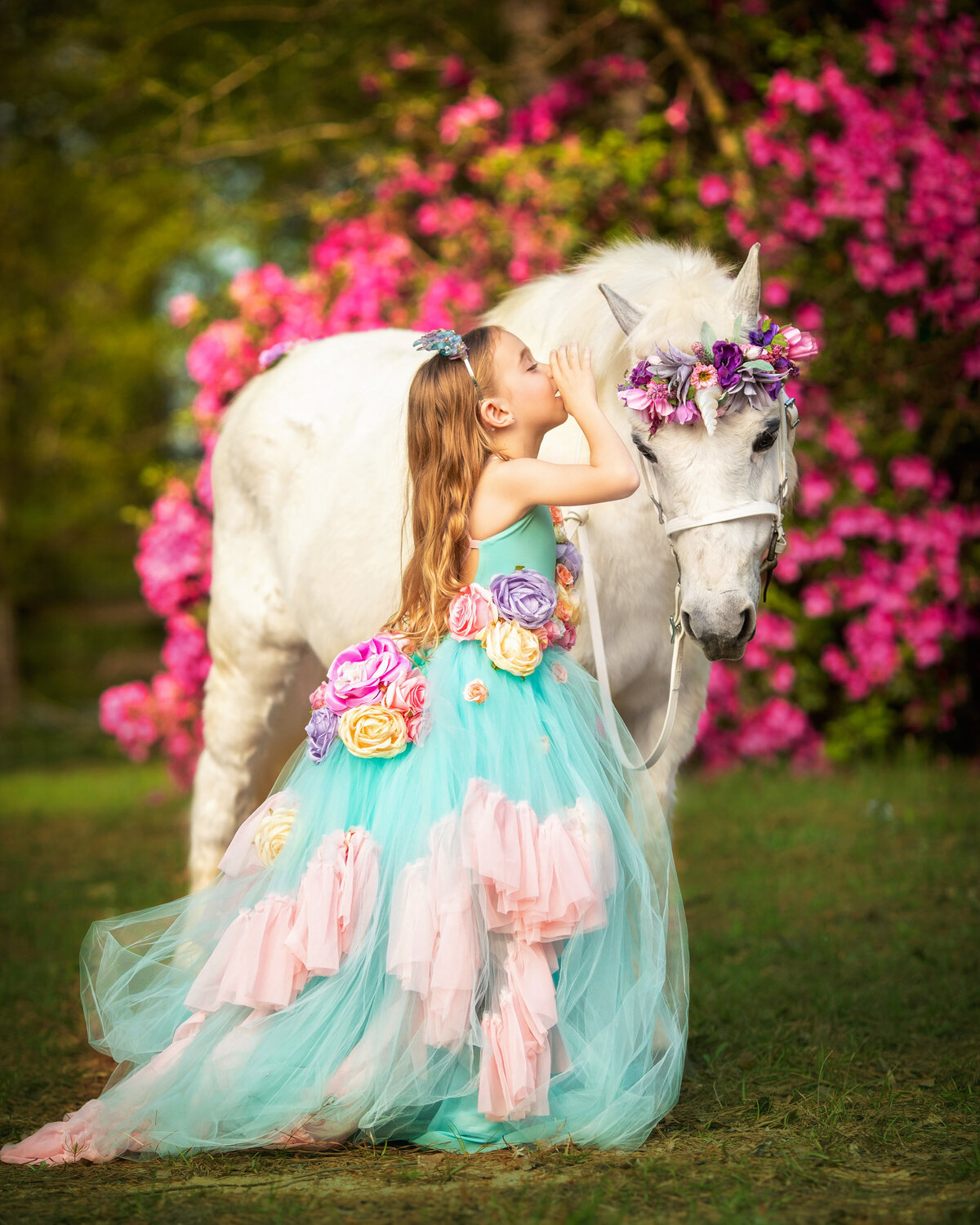 Welsh pony with a white unicorn horn and pink and purple flowers on her bridle.  The horse is white.  There is a young girl with long dark blonde hair.  She is wearing an aqua and pink dress with roses on it.  She is wispering into the unicorn's ear.  There are pink azaleals blooming in the background.