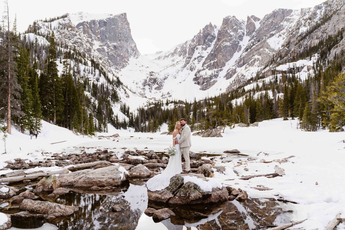 Couple stands on a rock beside the frozen waters of Dream Lake in Rocky Mountain National Park during their elopement.