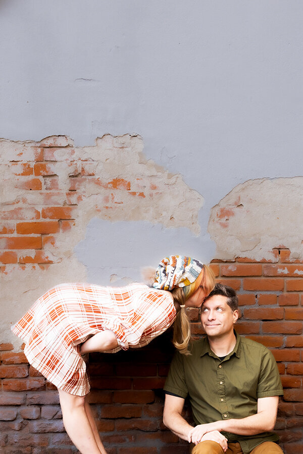 Girl kissing boy on head in front of brick wall.