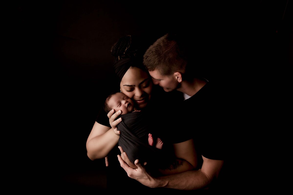 An intimate family photo from a newborn session with Mobile, AL Newborn Photographer. The family is all wearing black, posed on a black backdrop for a high-key photograph. The mom is holding her swaddled baby against her face, while dad wraps his arms around his wife and gently presses his face to hers to showcase emotion and connection.