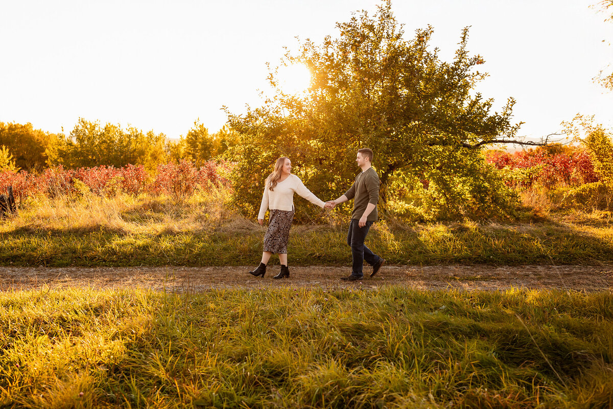 Women leading her fiancee along dirt road during fall sunset engagement session at apple orchard