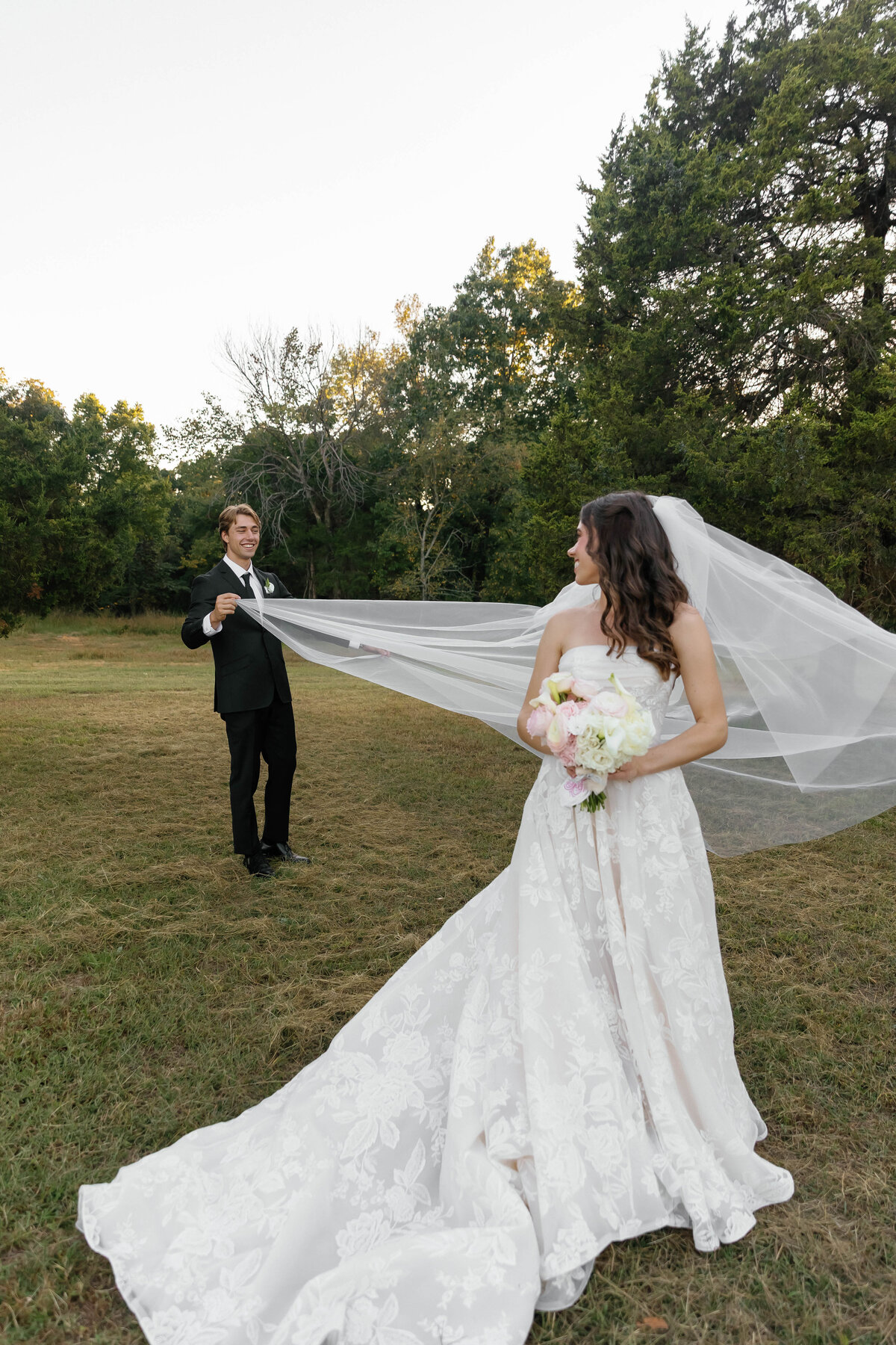 Bride and groom in an open field during sunset as the groom lifts and plays with the bride’s cathedral veil, highlighting the soft romantic florals in her bouquet styled by a fine-art florist.
