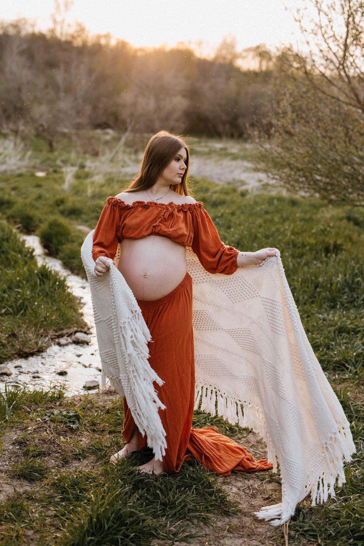 Mom standing in field in two-piece burnt orange top and skirt with belly exposed.  She is wrapped in a cream blanket flowing in the wind.