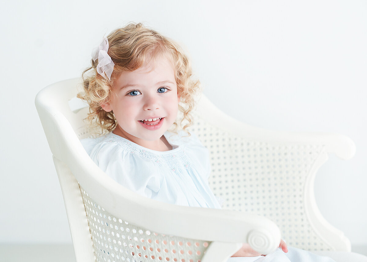 curly haired blond 3 year old photographed wearing a feltman brothers dress in a white wicker chair