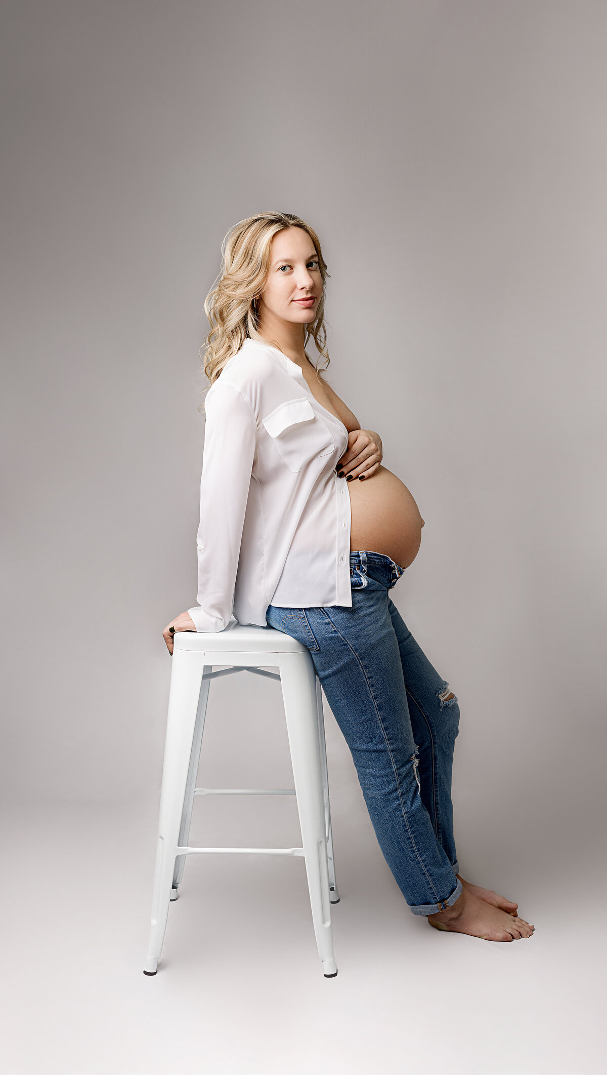 Mother wearing white button down with pregnant belly poking out leaning on white stoll