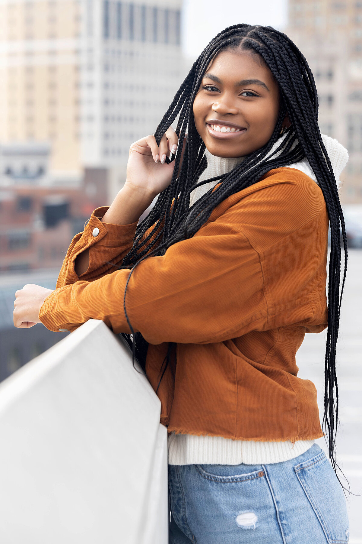Detroit Z Lot Garage Senior Photoshoot | Detroit Michigan Senior Photographer, Senior girl wearing dark tan corduroy jacket, white cowl neck sweater, and stonewash destructed jeans leaning on ledge overlooking downtown