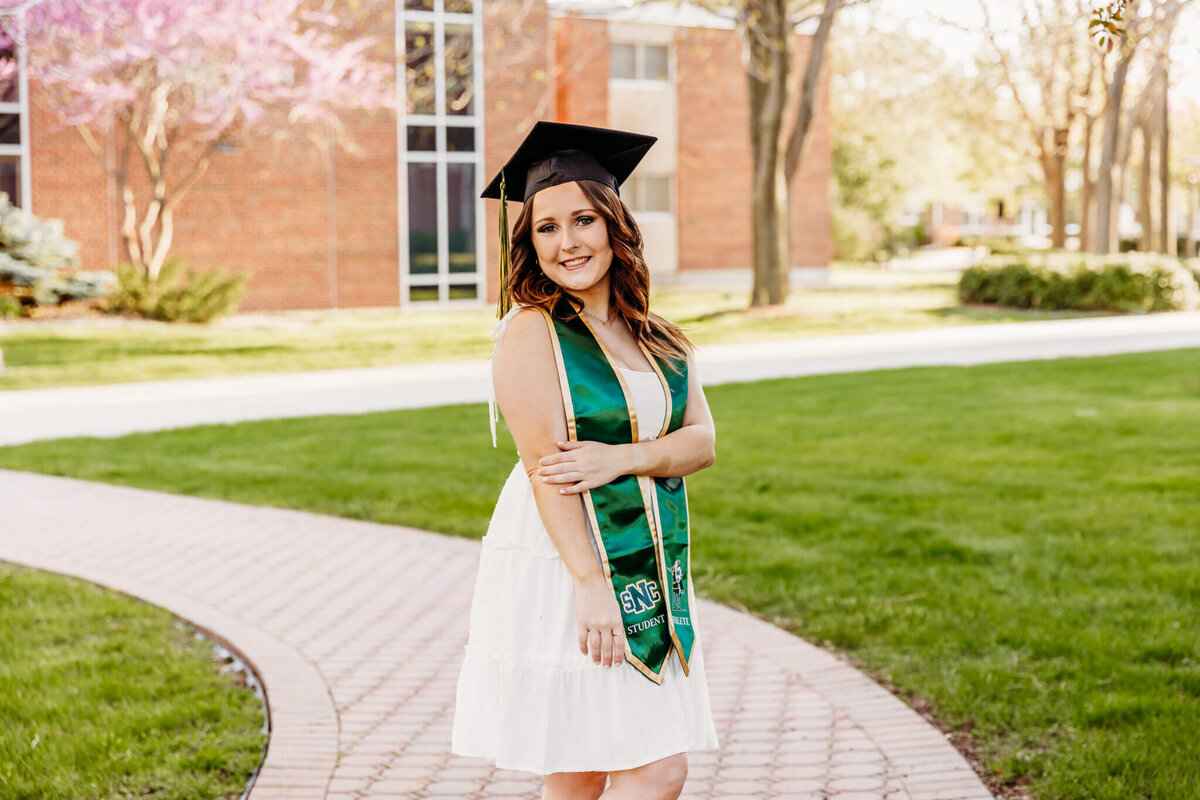 young woman wearing her grad cap and holding her elbow while smiling during her St Norbert graduation photo session
