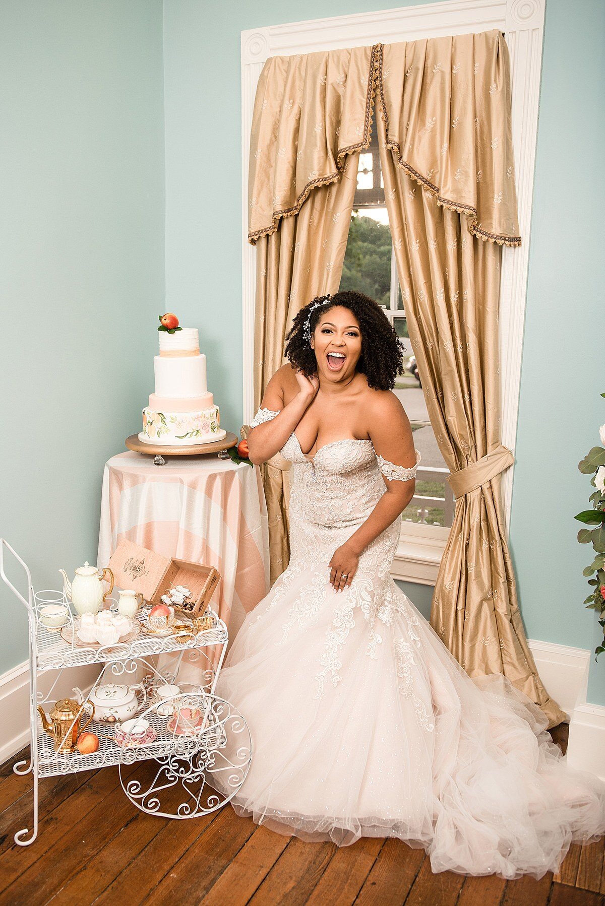 African American bride, wearing an off the shoulder lace mermaid wedding dress with a long train is very excited to see her five tier hand painted peach themed wedding cake and white metal tea and dessert cart at Ravenswood Mansion's blue room.