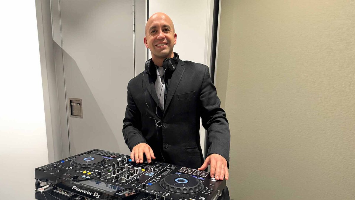 Male DJ in a black suit smiling behind a Pioneer DJ controller at a formal indoor event.