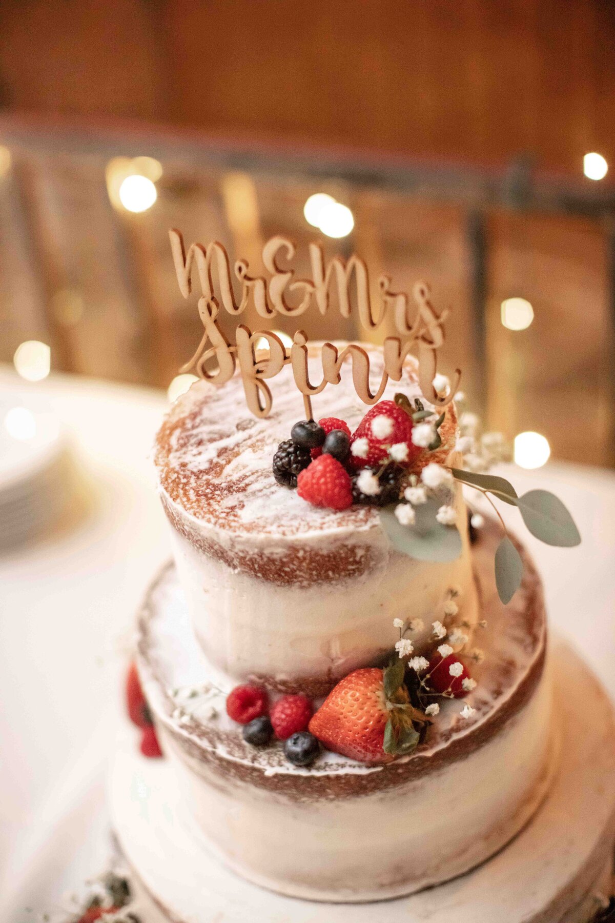 wedding cake with berries and twinkle lights behind wooden sign on top by Firefly Photography
