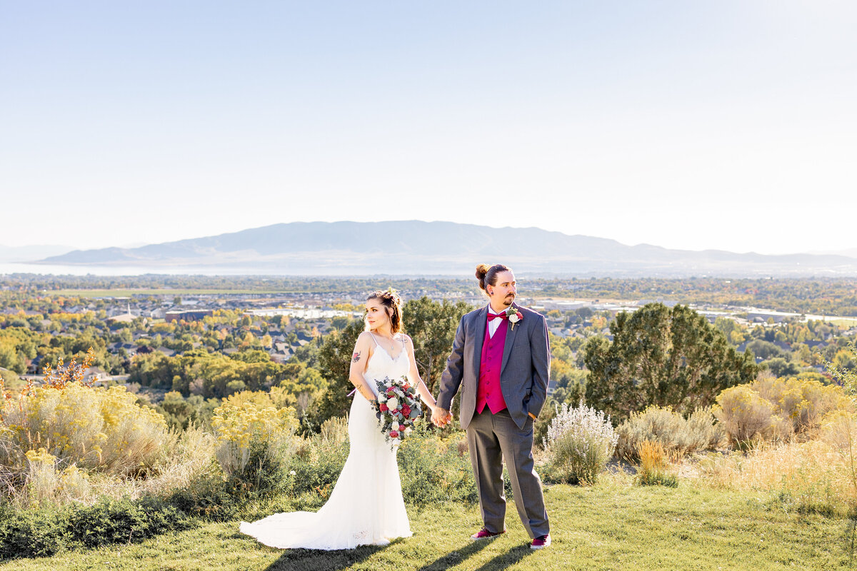 The bride and groom hold hands and look in opposite directions, posing in front of a stunning valley view with mountains in the distance and golden light illuminating the landscape in Highland, Utah.