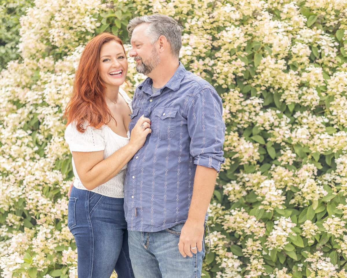 Husband and wife standing in front of a bush with white blooms.  Wife has a short sleeve shirt on and blue jeans with long red hair, smiling at the camera.  Husband is looking at her with a pepper hair and beard, wearing a button up shirt with the sleeves rolled up and jeans