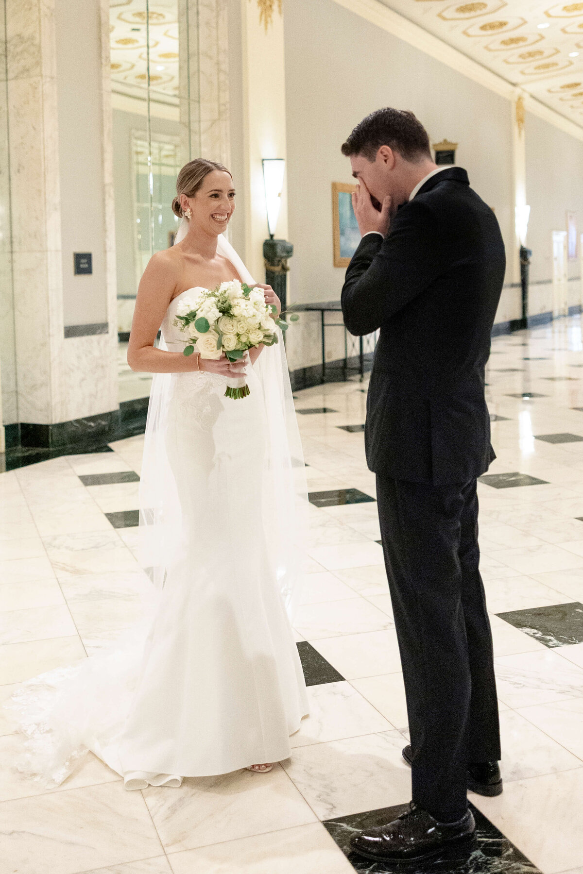 A bride in a white dress and veil holds a bouquet of white and pink flowers. She faces a groom in a black suit, who appears emotional, covering his face with his hands. They stand in an elegant, marble-floored hallway.