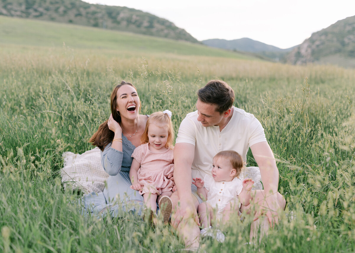 Family laying down and snuggling in a field holding their baby up in the air and smiliing at her- photo by Maegan R Photography