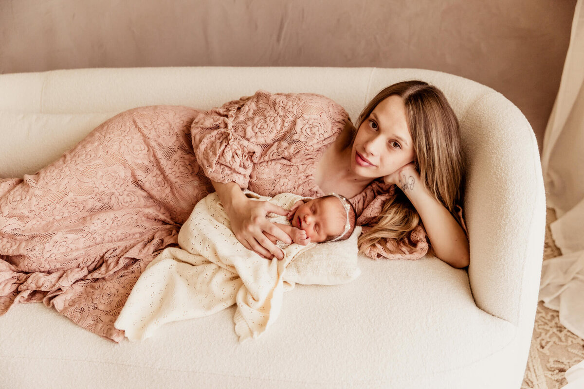 Mother laying on a white loveseat with her hand by her neck next to her newborn daughter.