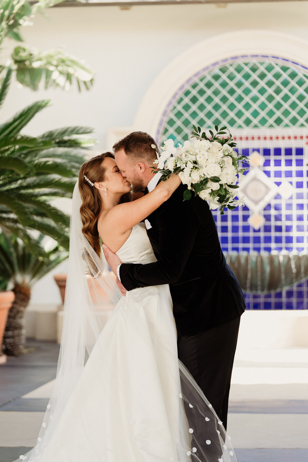 groom in a black tux  and bride in a white dress with a cathedral polka dot veil with a large white bouquet kissing at spanish hills country club captured by los angeles wedding photographer magnolia west photography