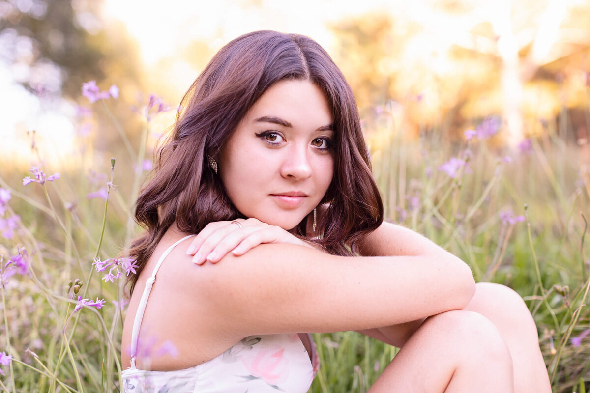 A woman with long brown hair sits in a field of purple flowers. She is wearing a light-colored, sleeveless dress and is looking at the camera with a calm expression. The background is softly blurred, highlighting the warm, natural setting.