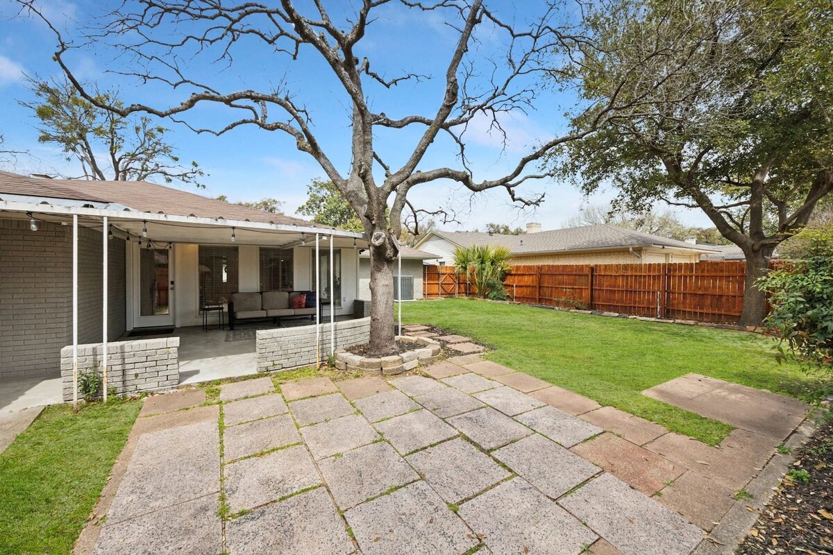 Backyard at 3120 Chapel Downs Dr Dallas TX featuring a covered patio, paved sitting area, and mature shade trees — Haven Realty Group.