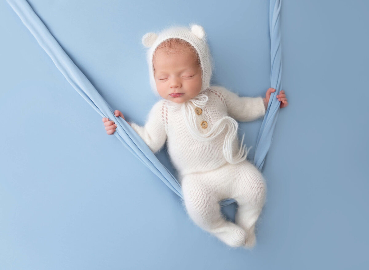 Newborn baby boy sleeping on a blue blanket wearing a white outfit with bear bonnet after the newborn pictures taken at NYC newborn photographer