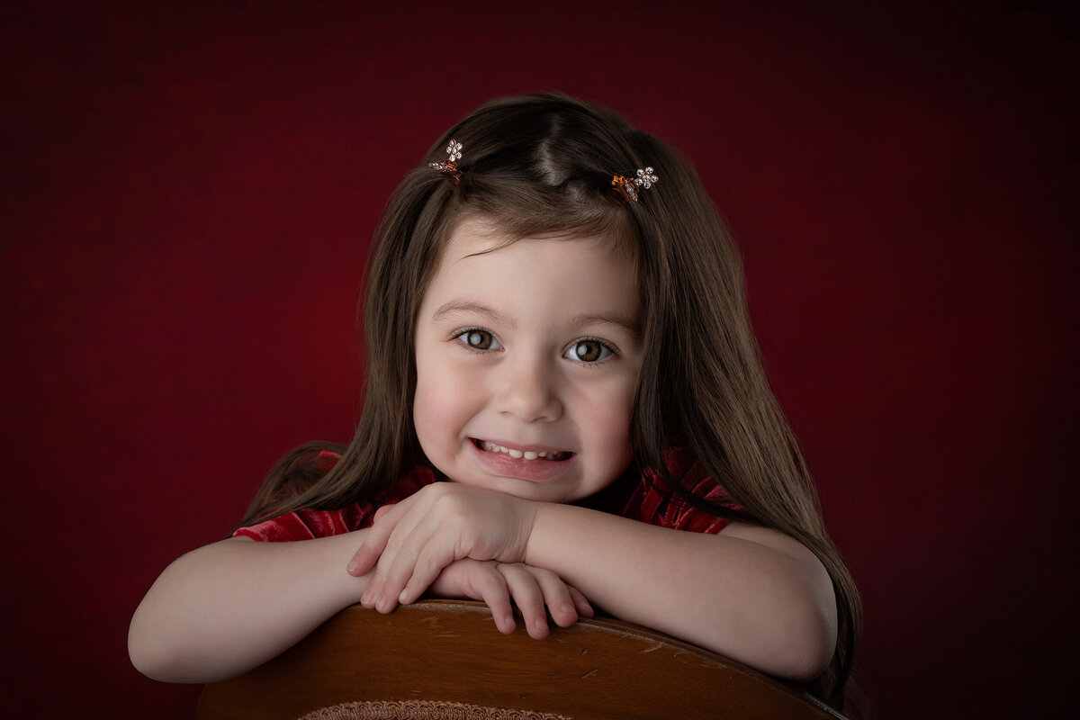 Young girl resting her chin on her hands on a chair, with a red background.