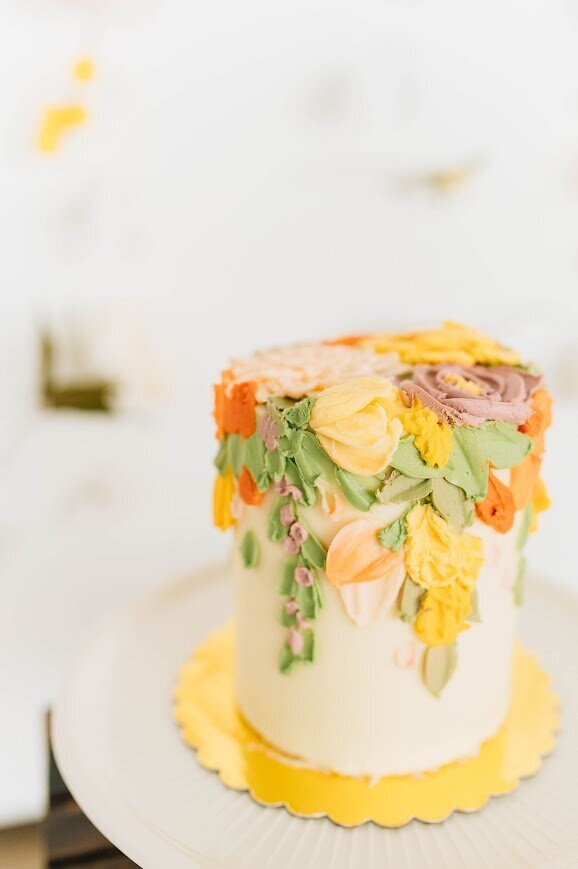 Photo is of the side of a cake. The base frosting is white, and the top of the cake is covered with buttercream painted florals in spring shades. The florals also come over and cascade down the sides of the cake.