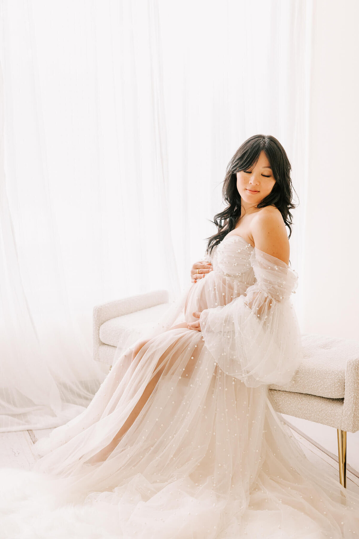 woman wearing pearl gown sitting in window looking over hear shoulder. She is pregnant and has her hands on her belly.