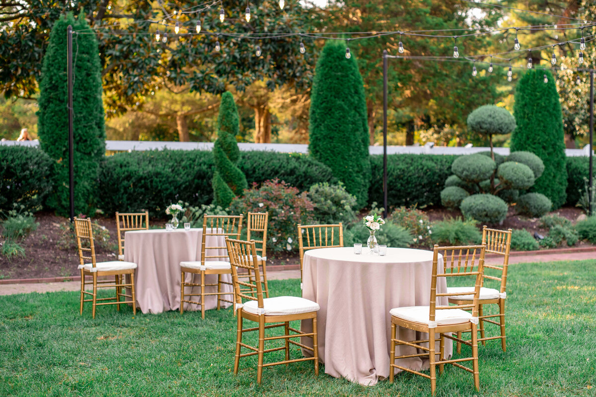 An outdoor setting with three round tables covered in light beige tablecloths, each surrounded by gold chairs. Each table holds small floral arrangements. The background features manicured hedges, topiaries, and string lights above.