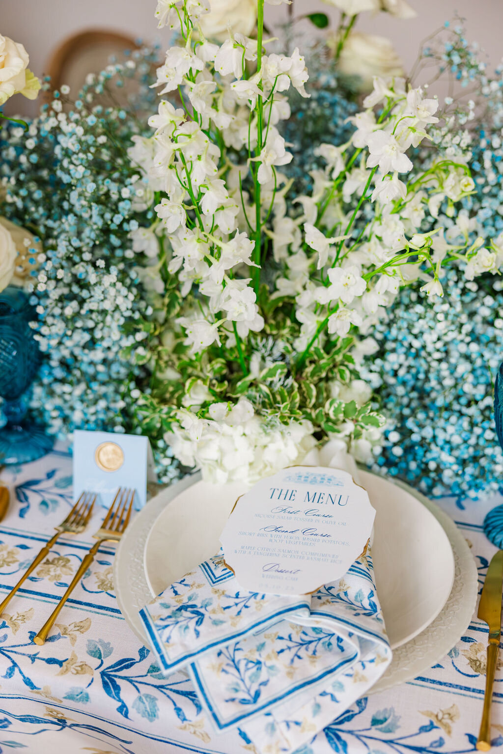 A detailed close-up of an elegant wedding place setting featuring a beautifully designed blue-themed menu card and coordinating napkin. The refined tableware, delicate floral accents, and sophisticated presentation create a timeless and luxurious dining experience, perfect for an upscale wedding reception.