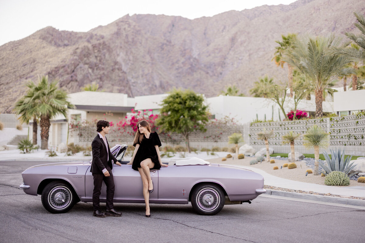 Stylish couple walking hand in hand beside a vintage lavender convertible with colorful mid-century buildings in the background.