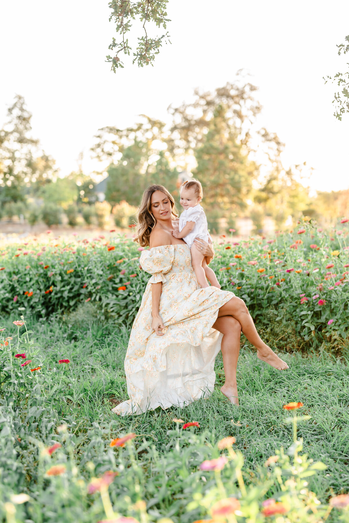 A mother with soft brown curls wearing a pale yellow floral gown sits in a field of zinnia flowers while holding her baby photographed by Bay Area Family Photographer, Light Livin Photography.