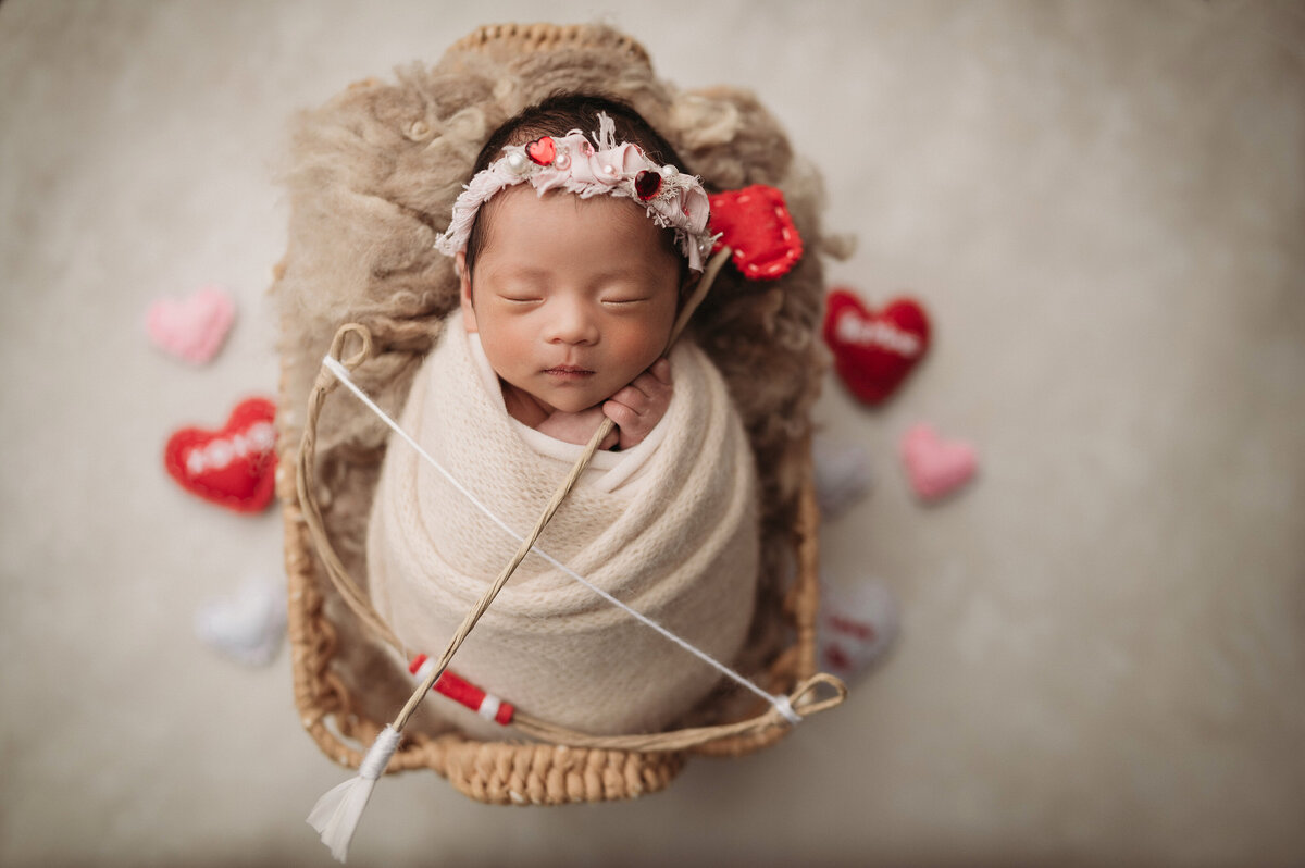 sleeping newborn baby girl  wrapped in cream swaddle and holding cupid's bow and arrow while laying in a layered rectagular wicker basket over a  cream background surrounded by valentine's day hearts