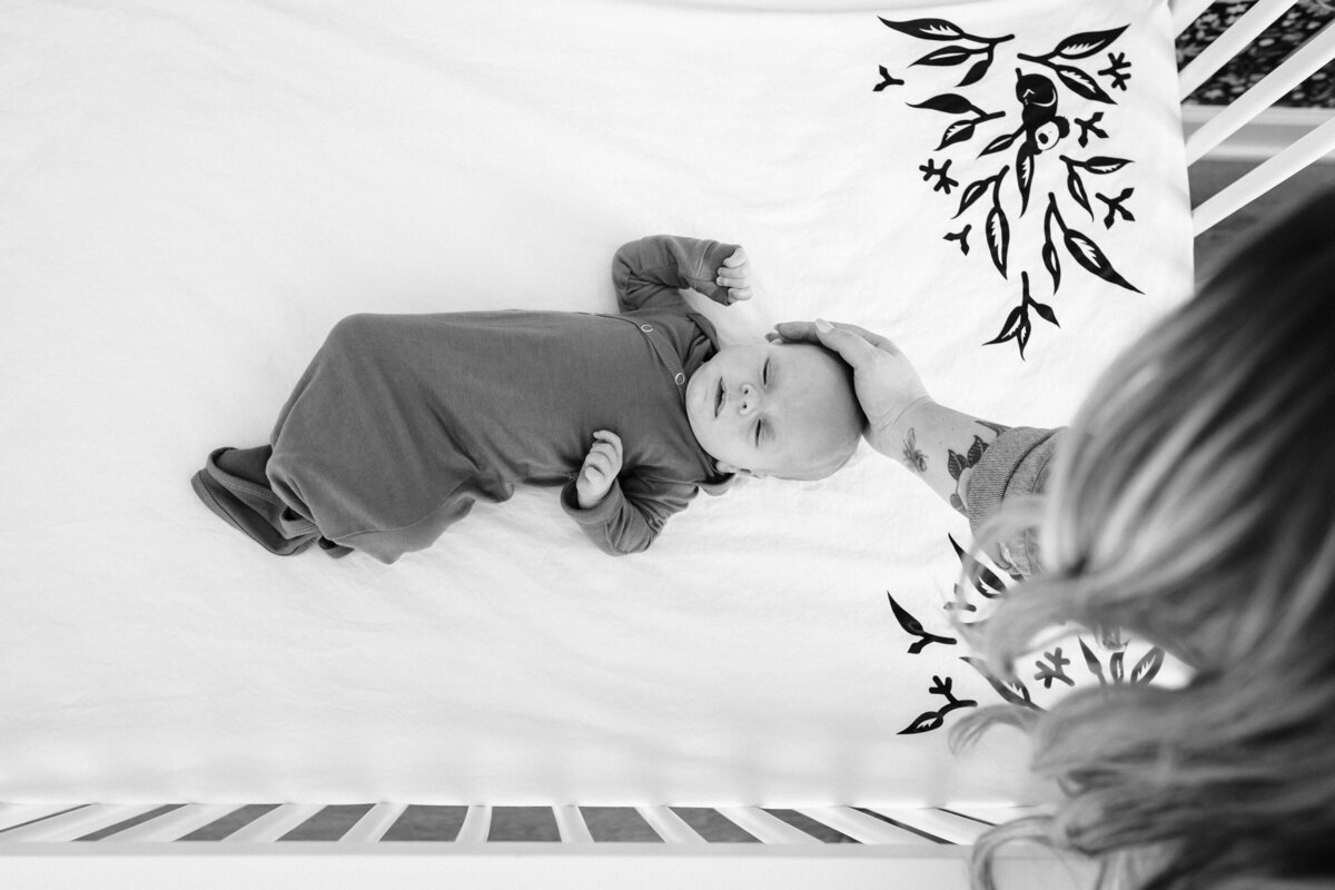 A black and white image of a mother gently placing her hand on her newborn's head as they lie swaddled in a crib with a floral patterned sheet.