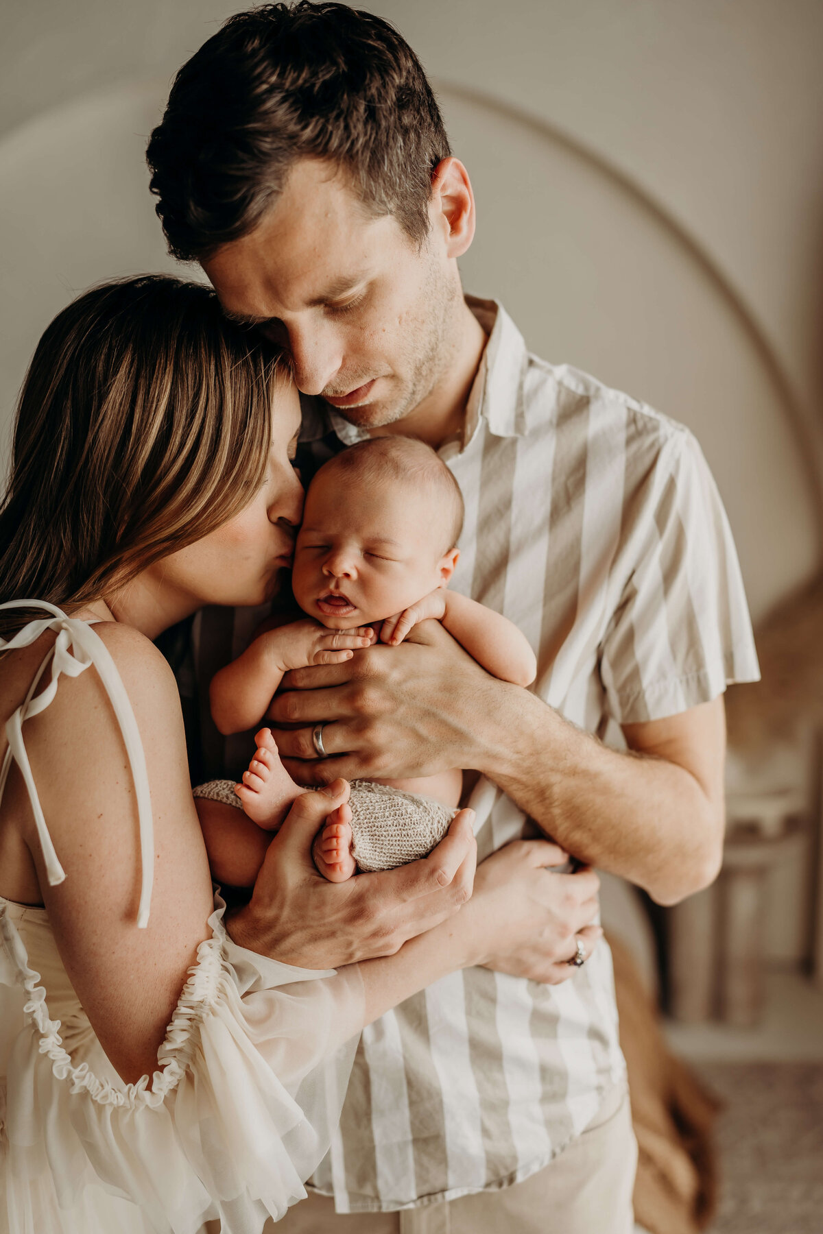 husband and wife hold their newborn and kiss him while wearing linen and chiffon wardrobe