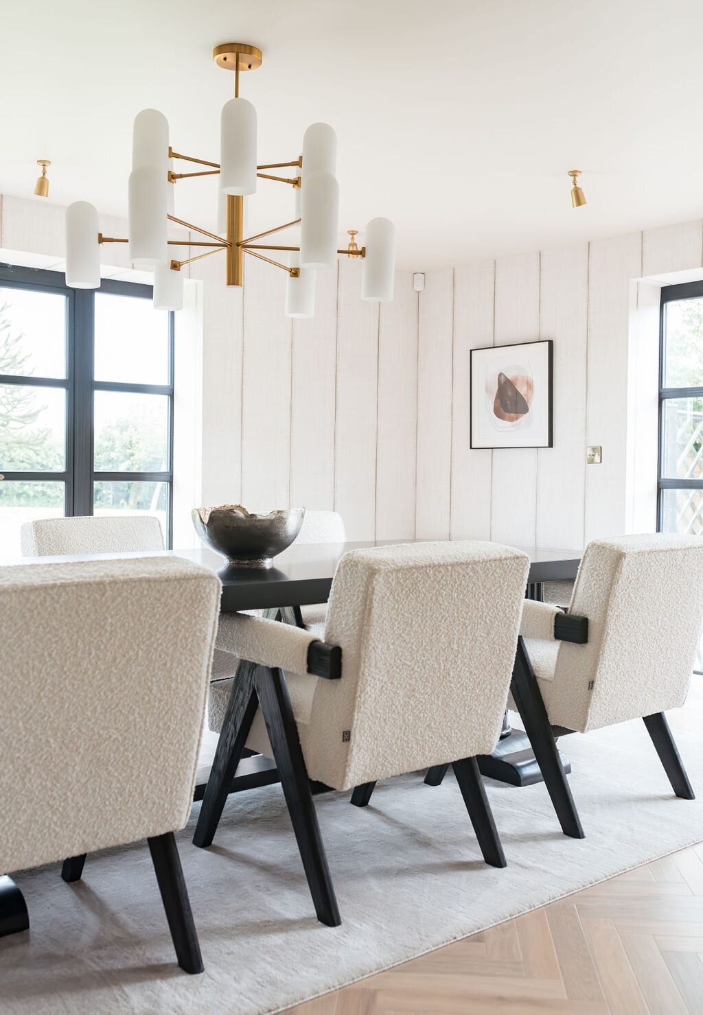 A modern dining room features a sleek black table surrounded by white upholstered chairs with black legs. A large white area rug covers the herringbone-patterned wooden floor. There is a gold chandelier and abundant natural light flooding through large windows.