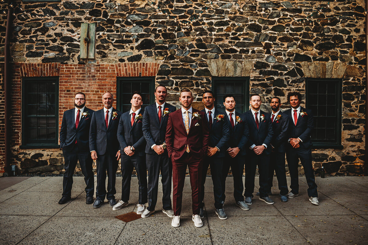 Baltimore wedding photographers captures groom in a burgundy suit standing with his groomsmen who are in gray suits outside of his Baltimore wedding venue