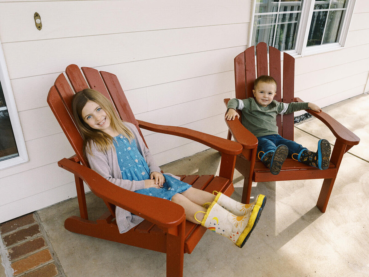 Two children sitting outdoors in red chairs during at home family photo shoot.