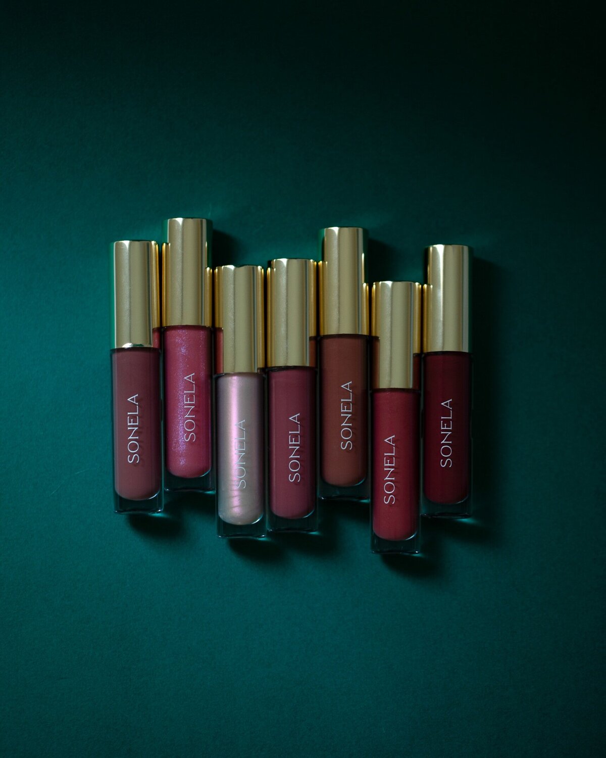 Sonela Beauty lip glosses with gold tops on a rich teal background