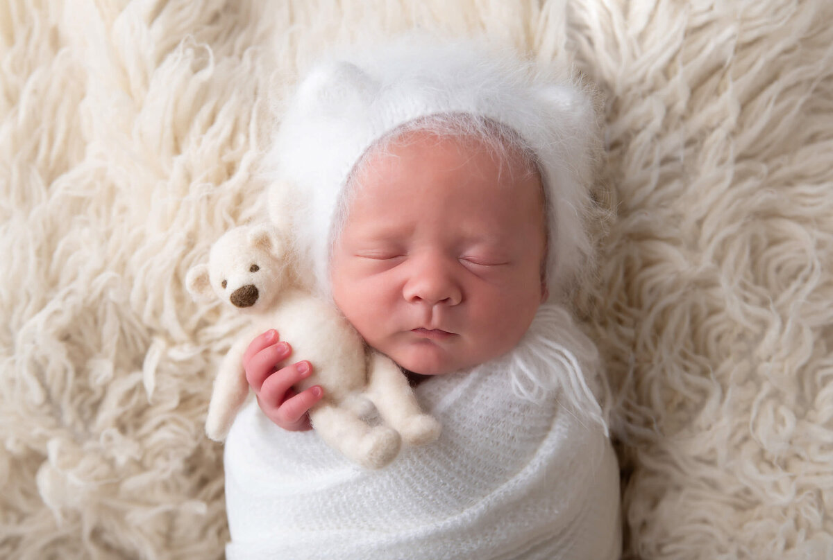 newborn baby boy wrapped in white, in a white bear hat , holding a white teddy for his newborn photos.