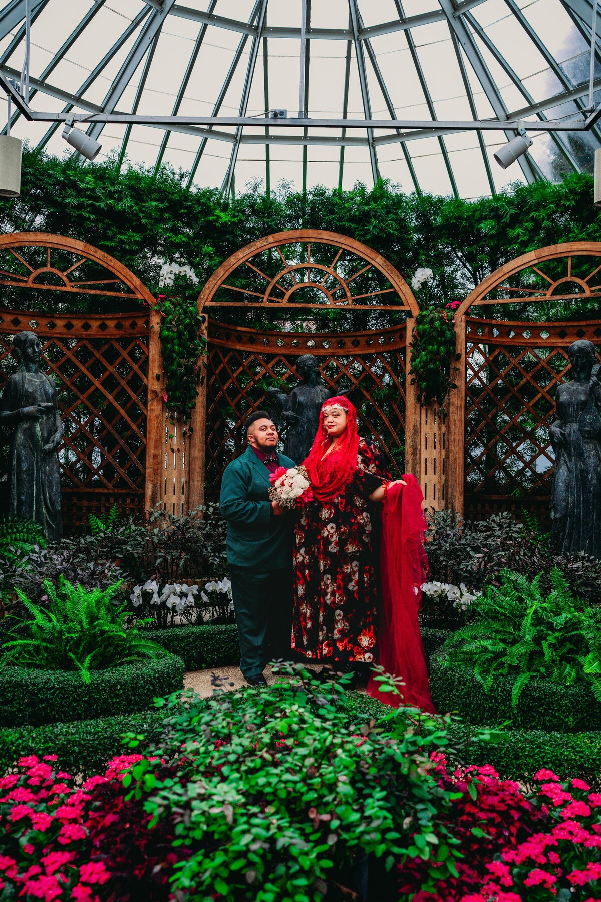 A queer couple shares a joyful wedding moment inside Phipps Conservatory in Pittsburgh, surrounded by vibrant flowers and lush greenery.