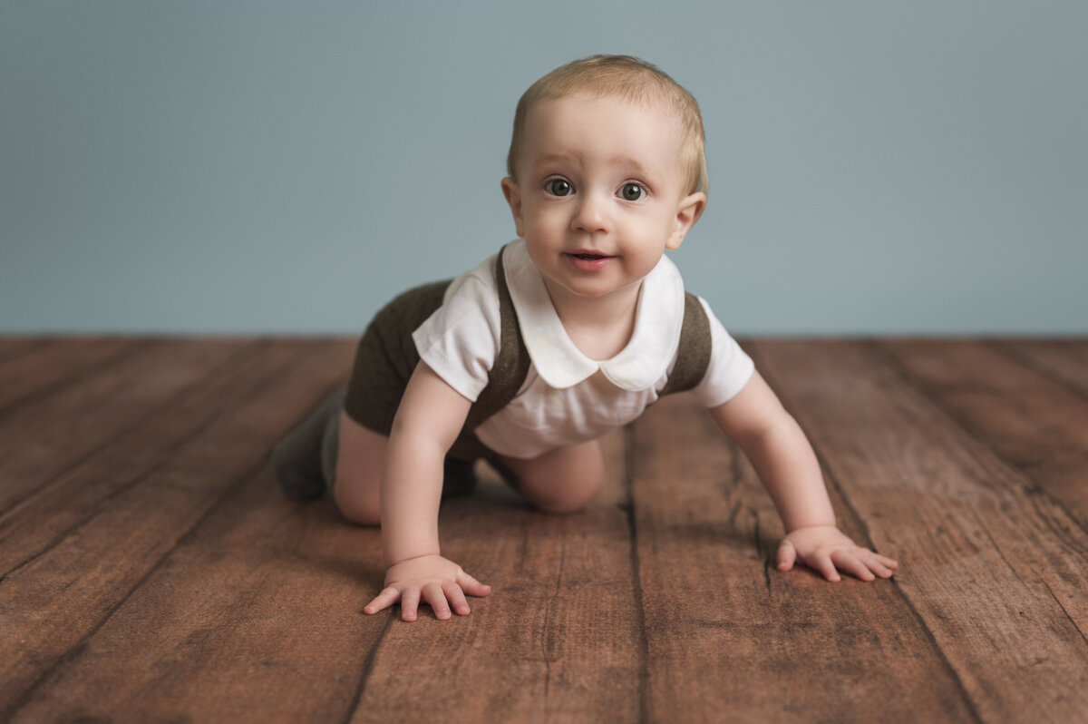 A crawling baby wearing a white shirt and brown shorts, smiling on a wooden floor with a blue background