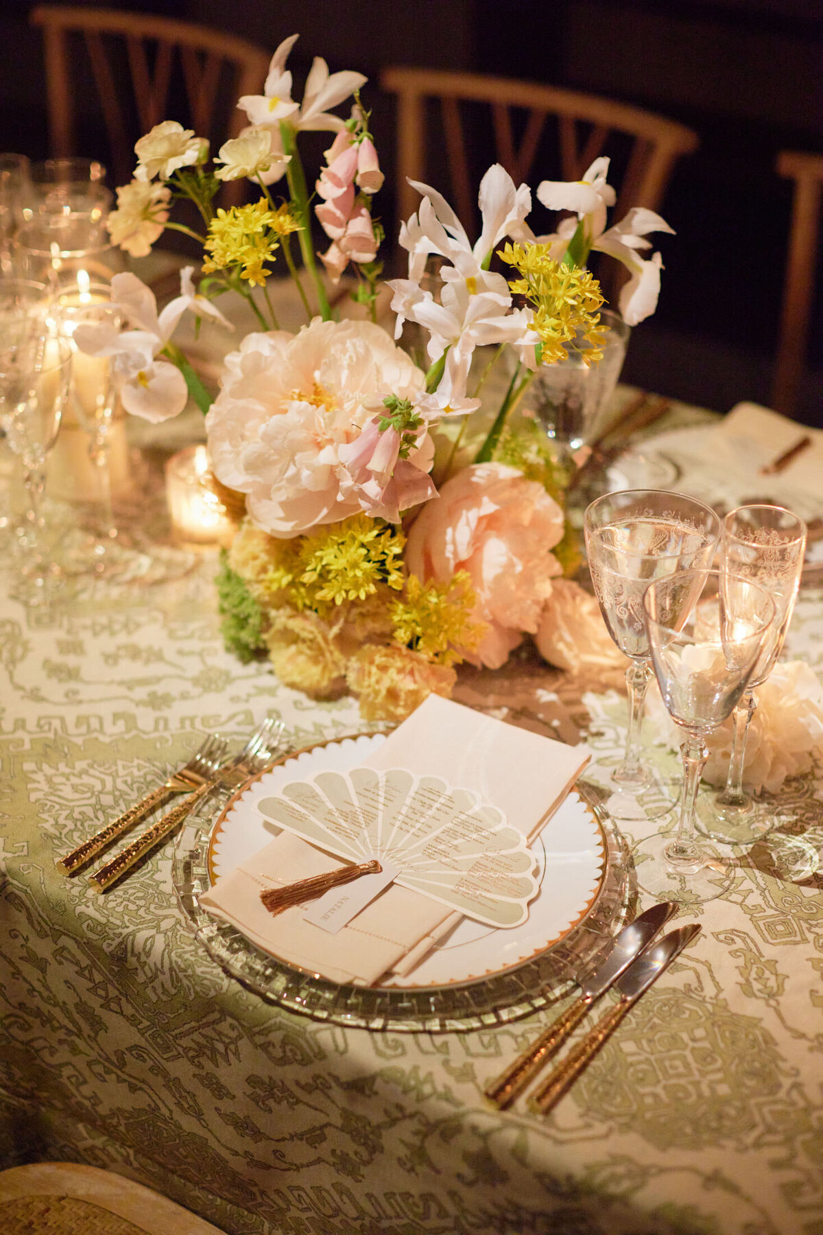 This is where the photo was taken at New York City, The New York Public Library by photographer Shawn Connell. Aaron Navak Films served as Videographers capturing the event. The beautifully set table features an elegant floral centerpiece with white and blush flowers, surrounded by candles. Each place setting includes a glass charger plate, a decorative fan-shaped menu, gold utensils, and crystal glasses on a patterned tablecloth.