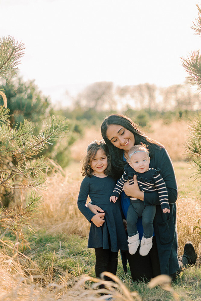 Mom holding her two young daughters at a Christmas tree farm