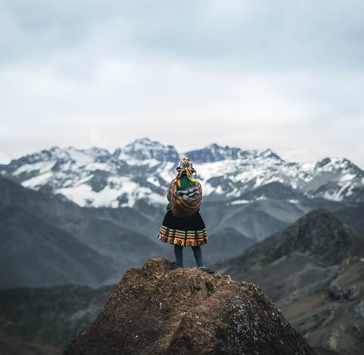 A traveler in traditional Andean attire stands atop a mountain peak, gazing at the snow-capped Peruvian Andes.