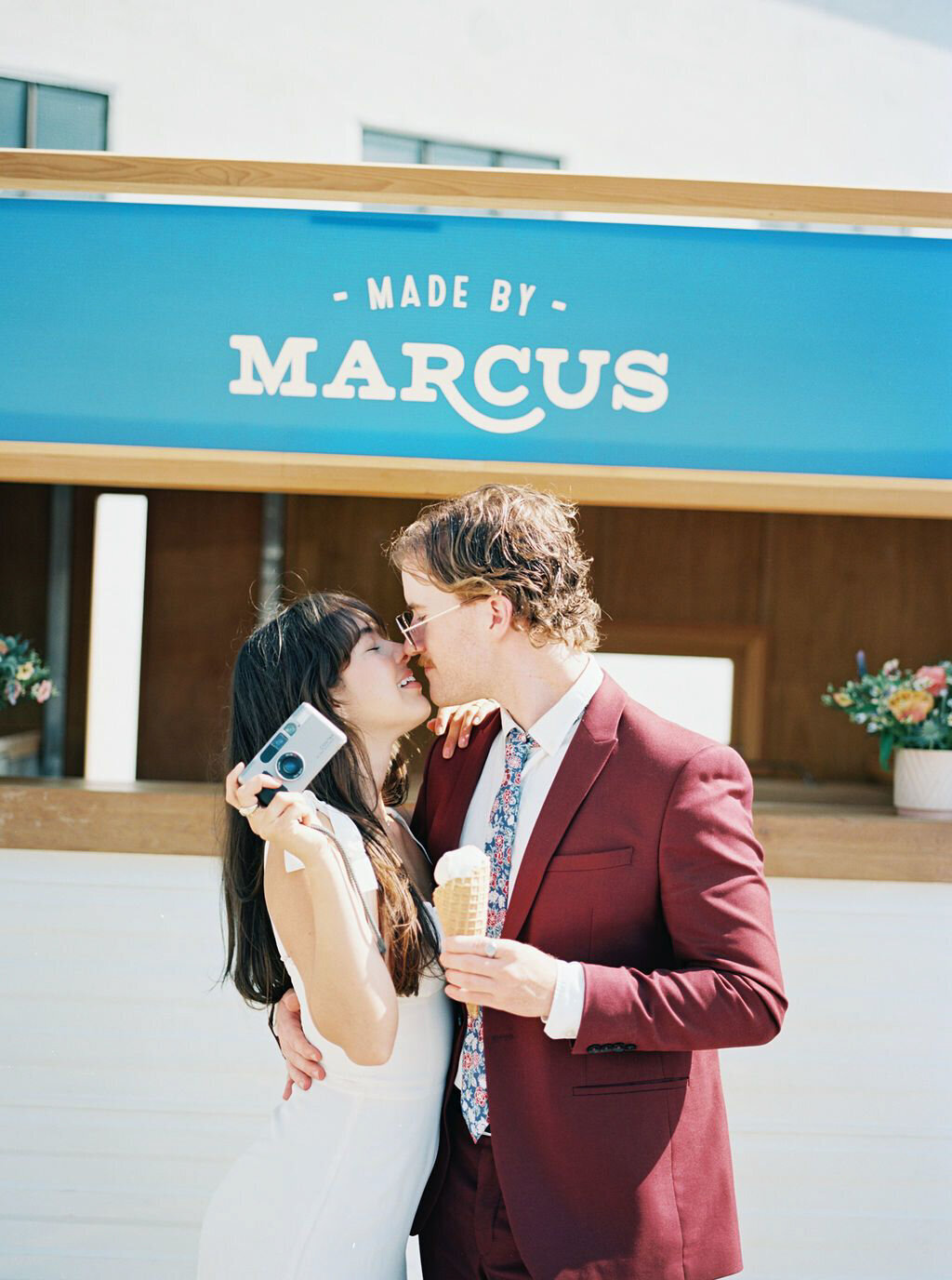 Groom wearing burgundy suit and floral tie, bride in sleek modern bridal gown in front of Made By Marcus, unique and playful ice cream based in Calgary, AB. Featured on the Brontë Bride Vendor Guide.