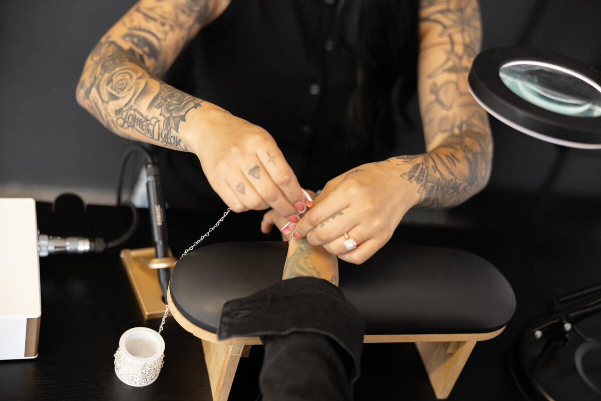 Tattooed woman with long dark hair adjusting tools while client rests in studio chair. Photograph by Yucaipa branding photographer Kaitlyn Dawn Photography.