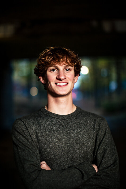 A close-up portrait of a smiling young man with wavy brown hair wearing a dark gray sweater. The background is dark with some soft bokeh lights creating depth.