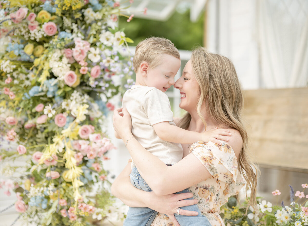 Mother smiling and embracing her son in front of a colorful floral arch during a spring greenhouse photo shoot.