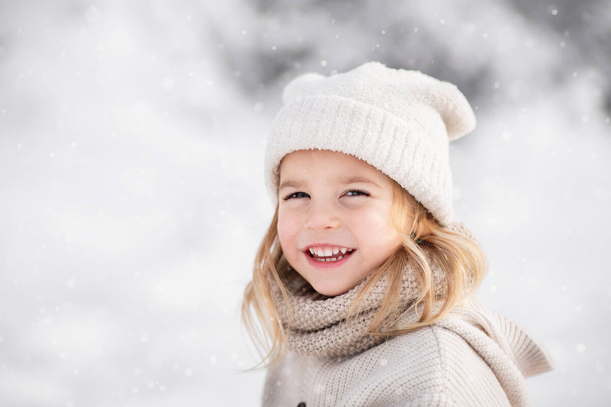 Brittany Danielle photographs a little girl with blonde hair wearing a white soft toque in the winter snow.