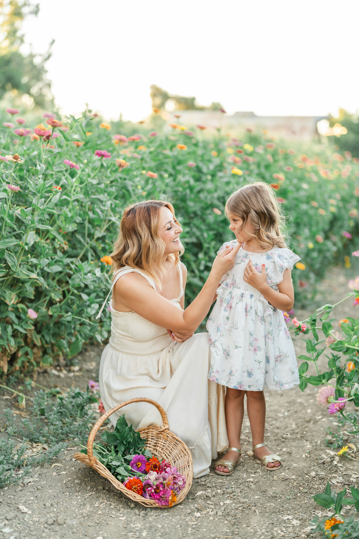 A mother with soft brown curls wearing a beige gown kneels and smiles next to her toddler daughter wearing a white and pink floral dress in a field of zinnias photographed by Bay Area Family Photographer, Light Livin Photography.